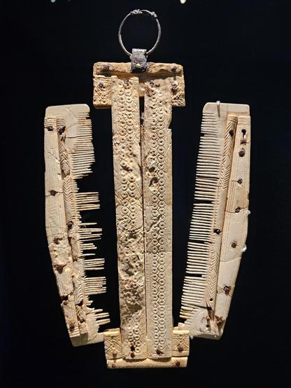 Three-part bone comb set: a narrow, ornamented central case with dense ring-and-dot decoration, flanked by two single-row combs with very fine, partly broken teeth, all hanging vertically from a simple round metal suspension ring against a dark background.