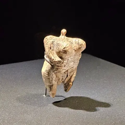 An ivory figurine with a weathered surface, shown in a museum setting, with exaggerated breasts, rounded hips, short limbs, and a small ring-like loop at the top where the head would be.