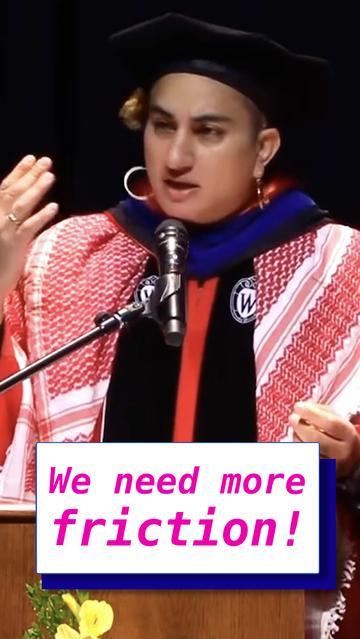 Alex, a brown trans woman with short curly black hair, is speaking into a microphone behind a wooden podium. She is wearing a red and black graduation gown, black graduation cap, and a blue hood around her neck. She is also wearing a red and white keffiyeh draped across her shoulders.
