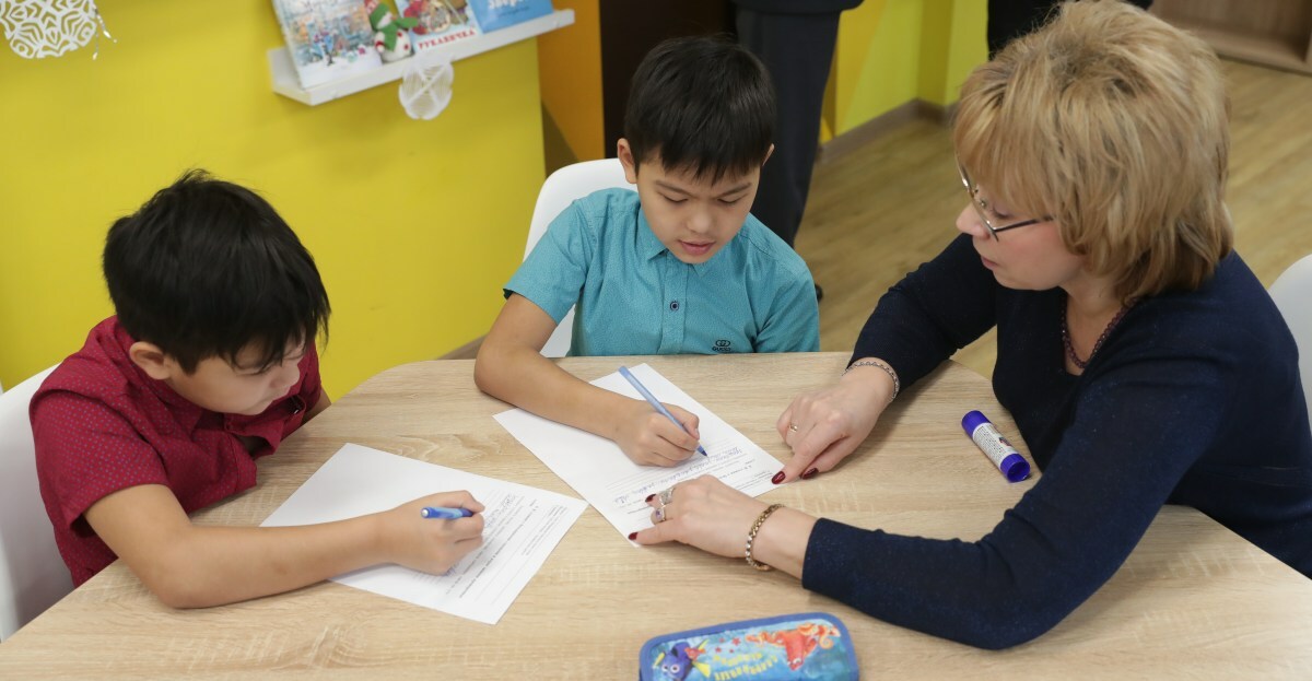 A teacher sits at a work table with two young students.
