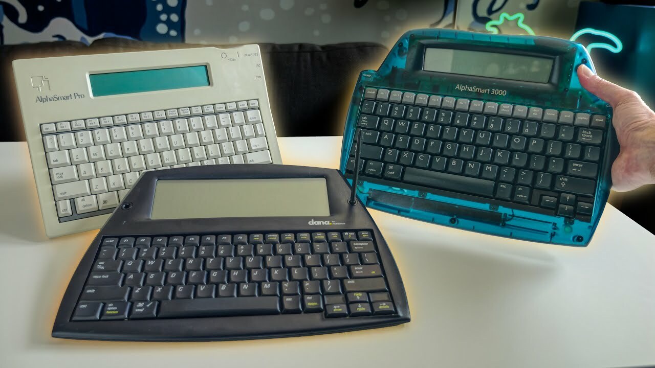 Three different keyboard devices with greyscale LCDs on top are on a white table. In the back left is a beige one with grey keys, front and center is a black model, and to the right is a translucent blue model with black keys being held up by a hand. Three different keyboard devices with greyscale LCDs on top are on a white table. In the back left is a beige one with grey keys, front and center is a black model, and to the right is a translucent blue model with black keys being held up by a hand.