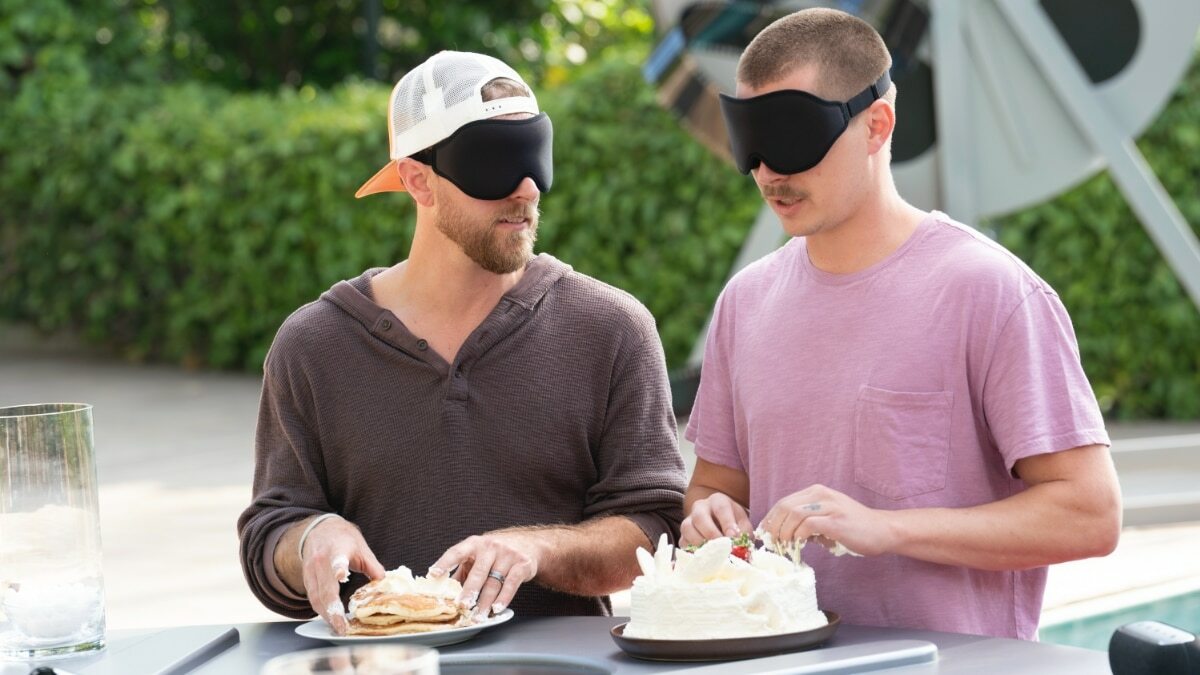 Two blindfolded people touch food in front of them: a stack of pancakes and a cake