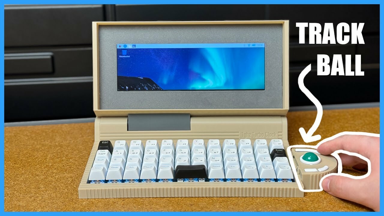 A very wide beige laptop sits on a wooden table. A hand manipulates a teal ball in a semicircle attached to the right sided of the device. The track ball and hand are outlined in white. A very wide beige laptop sits on a wooden table. A hand manipulates a teal ball in a semicircle attached to the right sided of the device. The track ball and hand are outlined in white.
