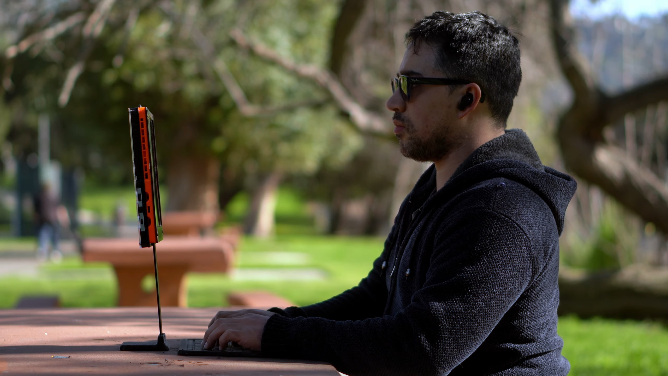 A man in glasses and a black sweatshirt sits in front of an orange and black computer screen just below eye level at the table in front of him. His keyboard sits on the table below. He appears to be in a park as there are trees and grass in the background. A man in glasses and a black sweatshirt sits in front of an orange and black computer screen just below eye level at the table in front of him. His keyboard sits on the table below. He appears to be in a park as there are trees and grass in the background.