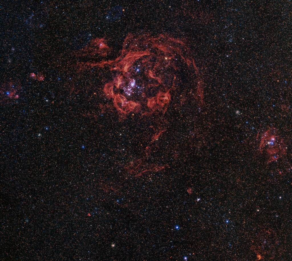 Thousands of distant stars crowd the view against black space. A rosy, bloomlike tendril of red nebulosity shines near the center-top.