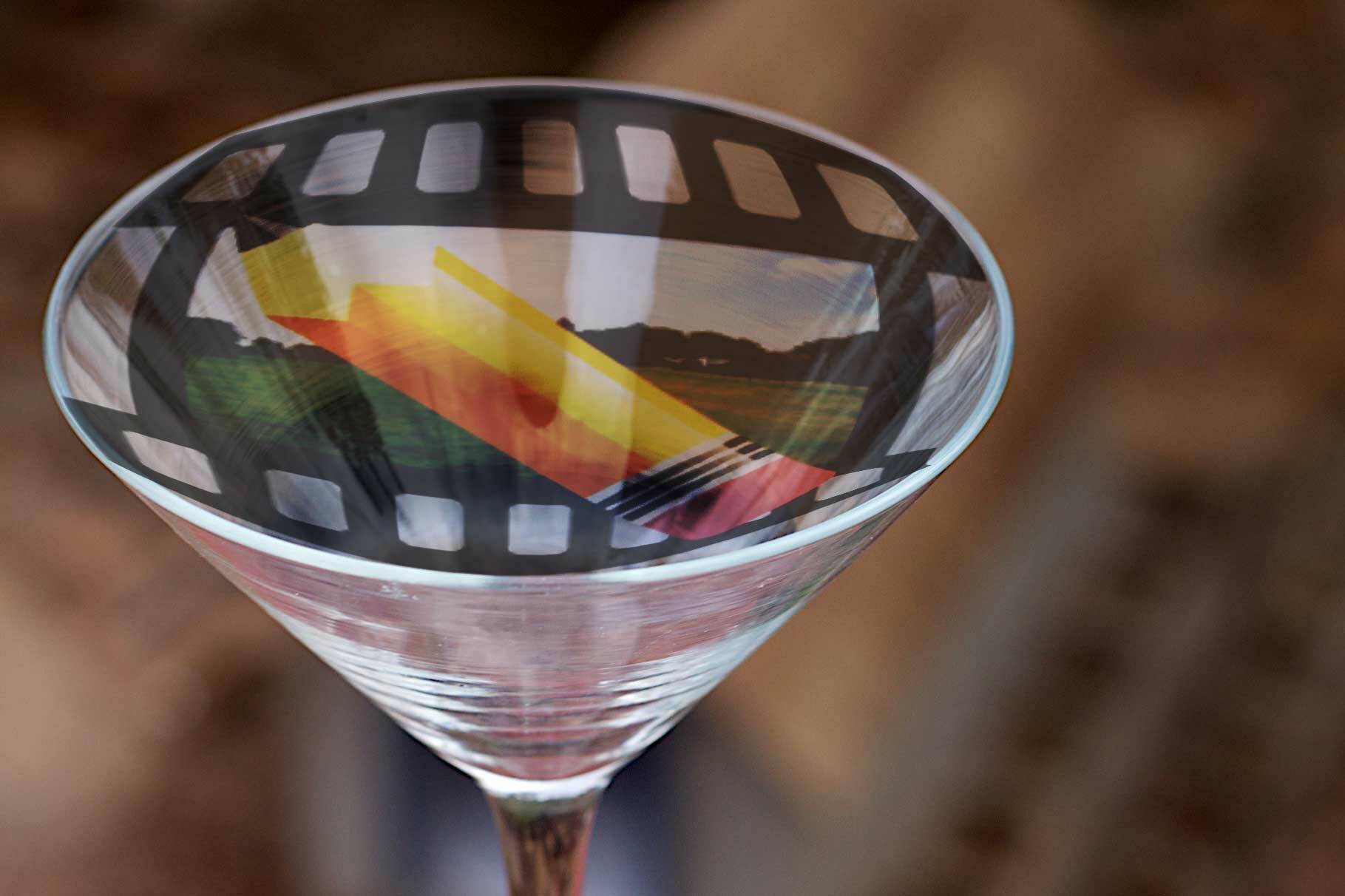 Photo of en empty martini glass, its convex/conical distinctinve shape sitting on a wood table, Superimposed on the op of the glass is the Photini logo, a camera film negative with a large pencil in the frame.