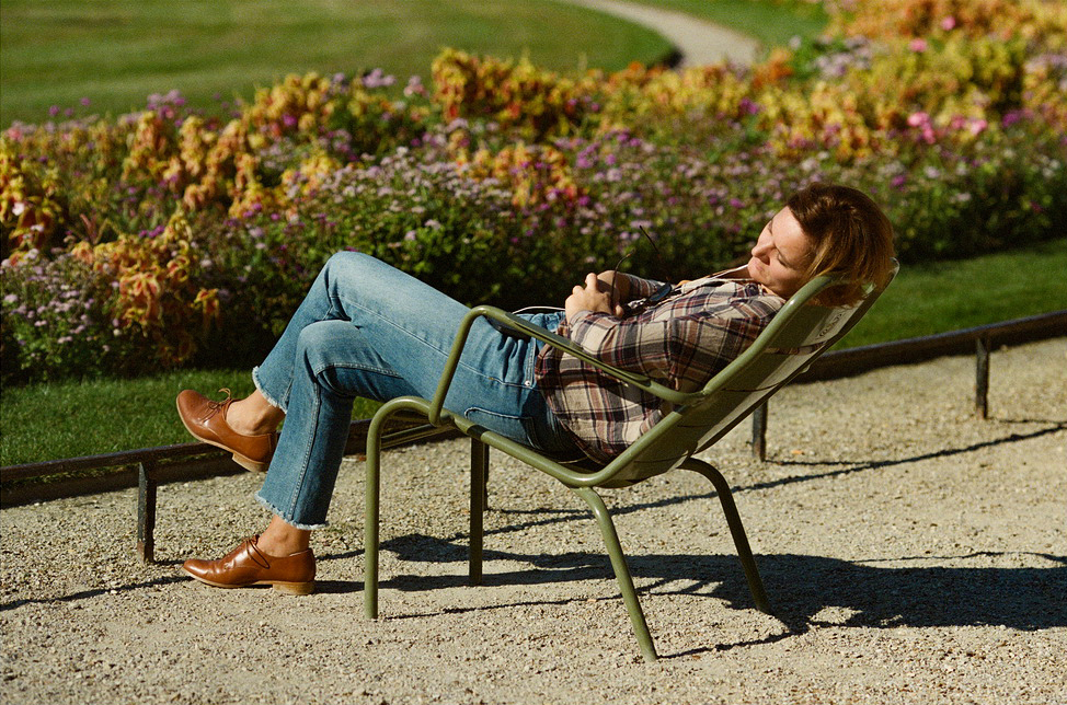 Caroline Berard, with short hair and wearing a plaid shirt and jeans, is reclining in a green metal chair outdoors. She appears to be asleep, arms folded across her chest. Behind her, there is a path and colorful flower beds in a sunny garden setting.