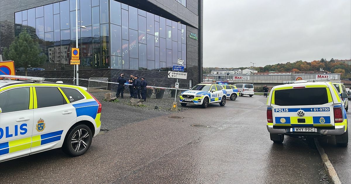 Polisbilar står parkerade vid en byggnad, där flera poliser diskuterar intill. Området är avspärrat med avspärrningsband och det är ett mulet väder.
