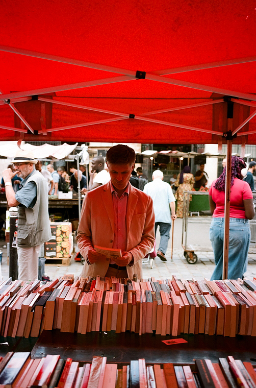 A person stands under a red canopy at an outdoor market, browsing through a table full of books. Several people are seen in the background, walking by other stalls and engaging in various activities. The lively and vibrant scene captures the essence of Ismael Lavaud's local community events.