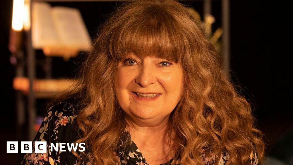 Janey Godley looks into the camera smiling. She has long curly brown hair with a fringe and is wearing a black floral blouse