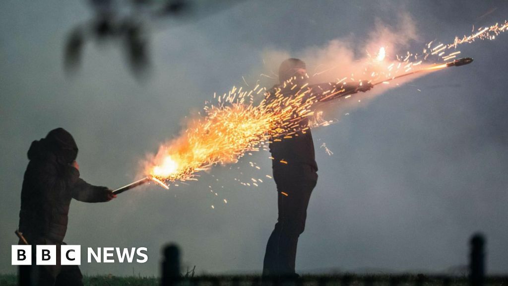 Two masked youths weaing black trousers and hoodies set off fireworks from their hands, sparks fly and the rockets launch in a smokey night time scene