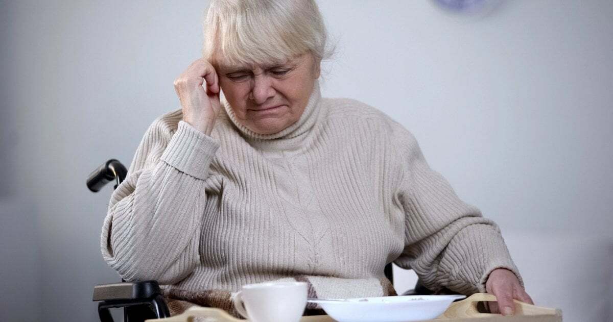 Eine ältere Frau, im Rollstuhl sitzend, blickt angewidert auf ein Tablett mit Essen.