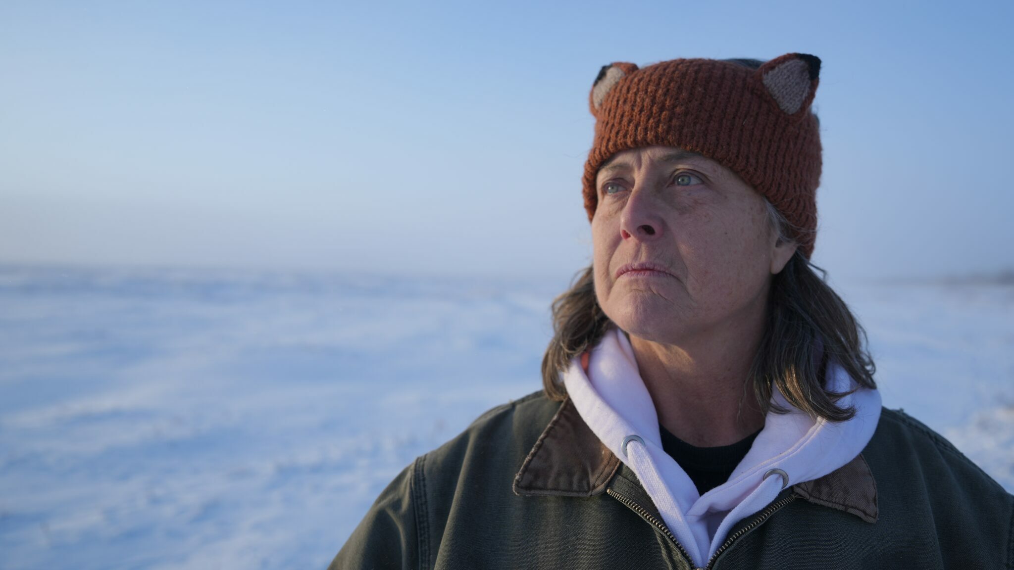 A person wearing a winter hat with animal ears looks off into the distance; snow is behind her