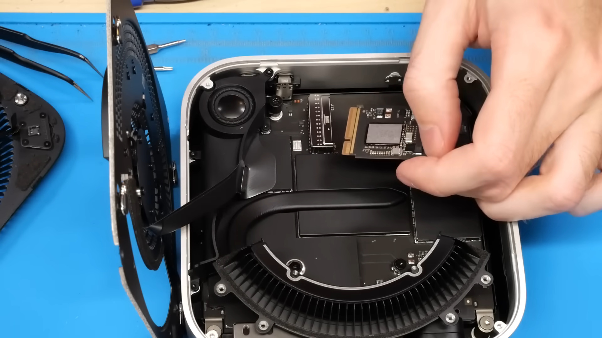 A hand holds a small PCB with an edge connector over the exposed, mostly black components of an M4 Mac mini. The bottom cover is hanging by an FFC cable off to the left of the A hand holds a small PCB with an edge connector over the exposed, mostly black components of an M4 Mac mini. The bottom cover is hanging by an FFC cable off to the left of the