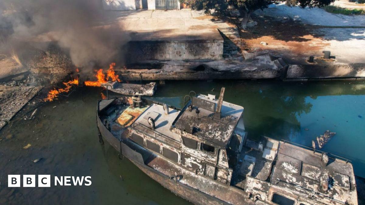 A burnt boat sits in the water with a fire burning close to it at the port city of Latakia on 10 December 