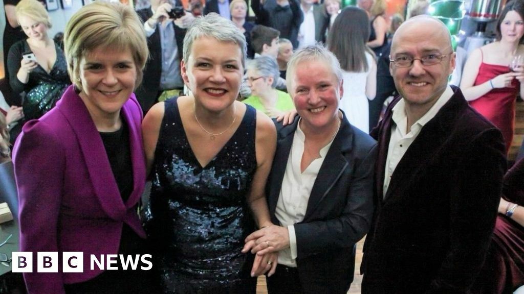 Nicola Sturgeon in purple jacket and dark blouse, Susan Douglas-Scott in a blue dress, Gerrie Douglas-Scott in a dark blue suit with white shirt and Patrick HArvie in purple velvet jacket are standing left to right at a party, there is a crowd milling about behind them. All four are smiling at camera.