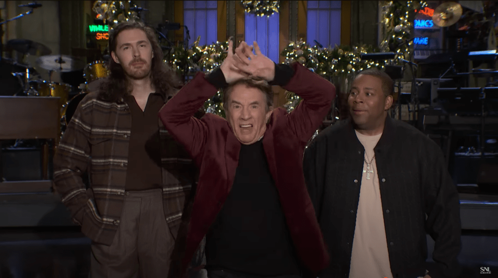 Martin Short looks angry with his hands above his head as he stands with Hozier and Kenan Thompson on a Christmas-decorated stage in a promo for 'Saturday Night Live'.