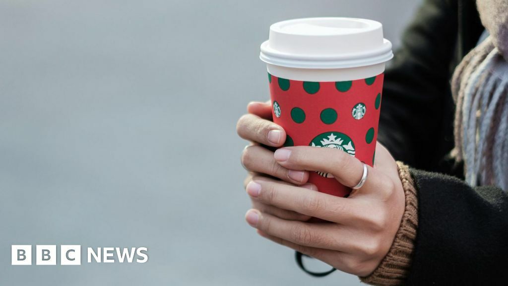 A woman holds a Christmas-themed Starbucks coffee cup. She's wearing a black coat, scarf and a silver ring on her left index finger.