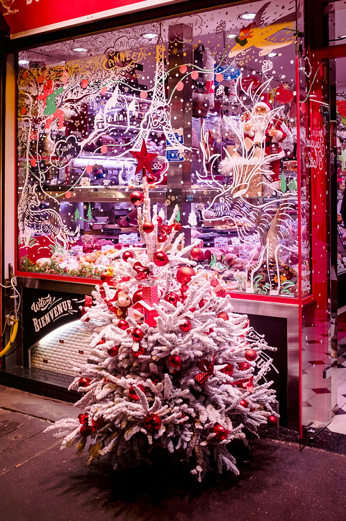 A small white Christmas tree adorned with red ornaments and a star topper stands in front of a store window. The window, brimming with Noël spirit, features festive illustrations full of snowflakes, colorful graphics, and a warmly lit interior displaying various decorations and products.