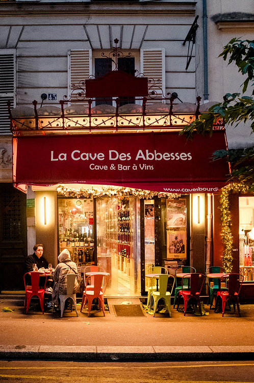 Cozy wine bar with a bright red awning labeled "La Cave Des Abbesses." Warm lights glow around the entrance, with colorful chairs at small tables. Two people are seated outside, creating a welcoming ambiance in the evening.