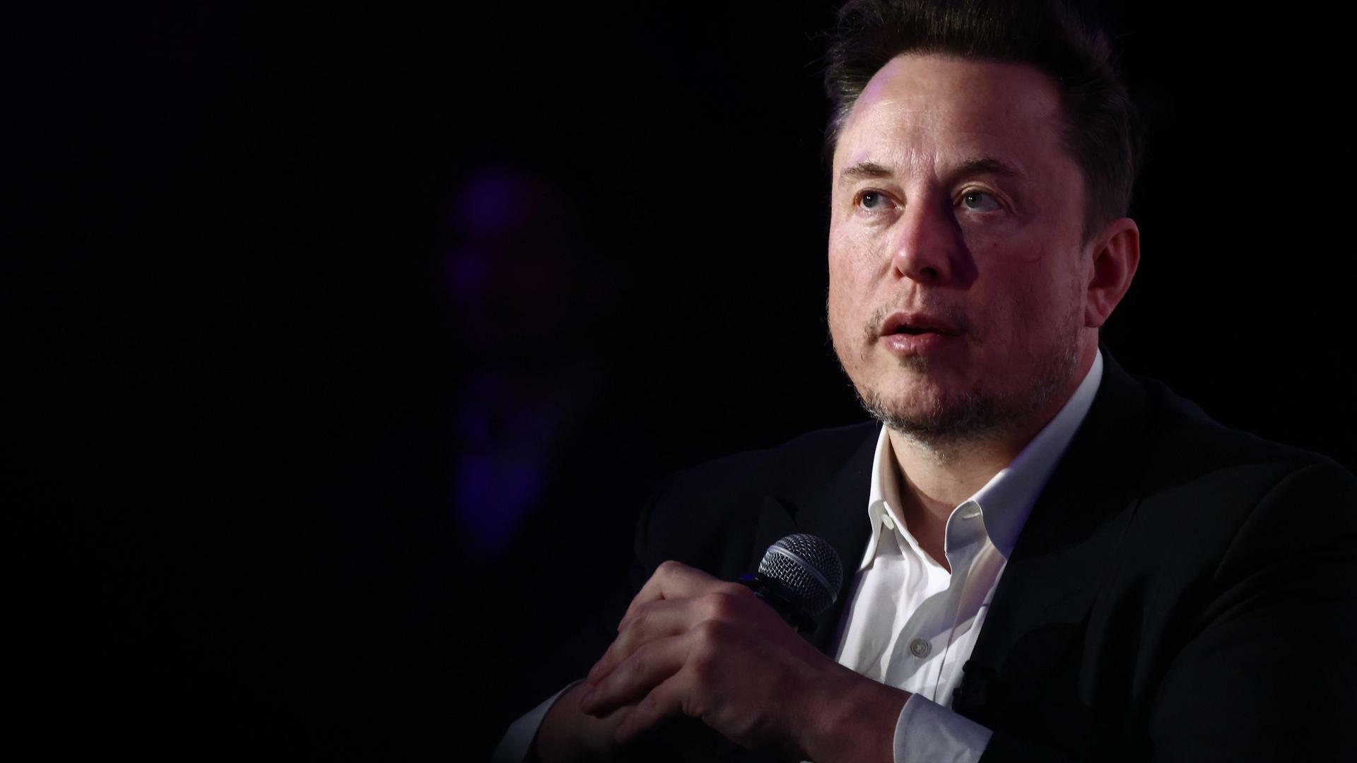 Elon Musk trägt einen Anzug und sitzt vor einem dunklen Hintergrund, in den Händen hält er ein Mikrofon.