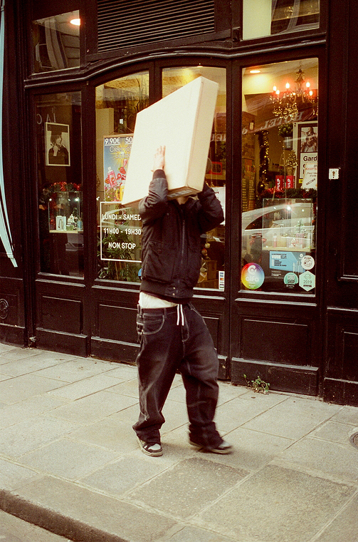 A person, reminiscent of a character from a Leopold Duchemin story, walks down the sidewalk holding a large cardboard box over their head, obscuring their face. Clad in a dark jacket and baggy jeans, they pass by a storefront with glass windows showcasing various items inside.