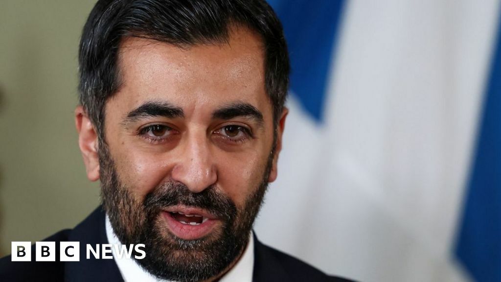 Humza Yousaf, with dark hair and a dark beard, photographed in a close-up shot with this mouth open as he speaks. A saltire is hanging on a wall behind him.