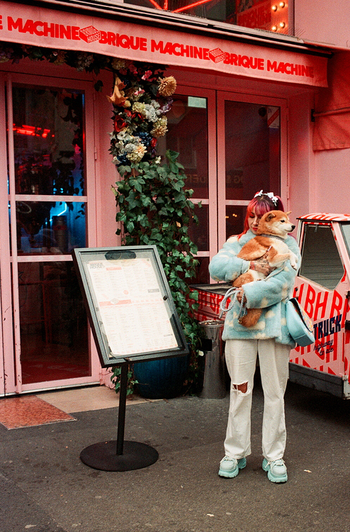Cosette Poirier, with her striking pink hair, stands outside a charming pink storefront holding a dog. Dressed in a blue jacket and white pants, she complements the scene's lively vibe. Nearby, a menu board adds character, while plants and red neon accents complete the picturesque setting.