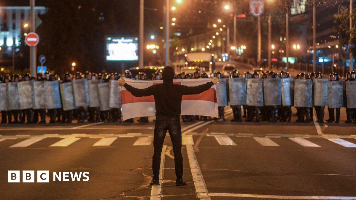 A protest is Belarus, August 2020. A lone man holds a white flag with a red stripe up to a crowd of riot police. 