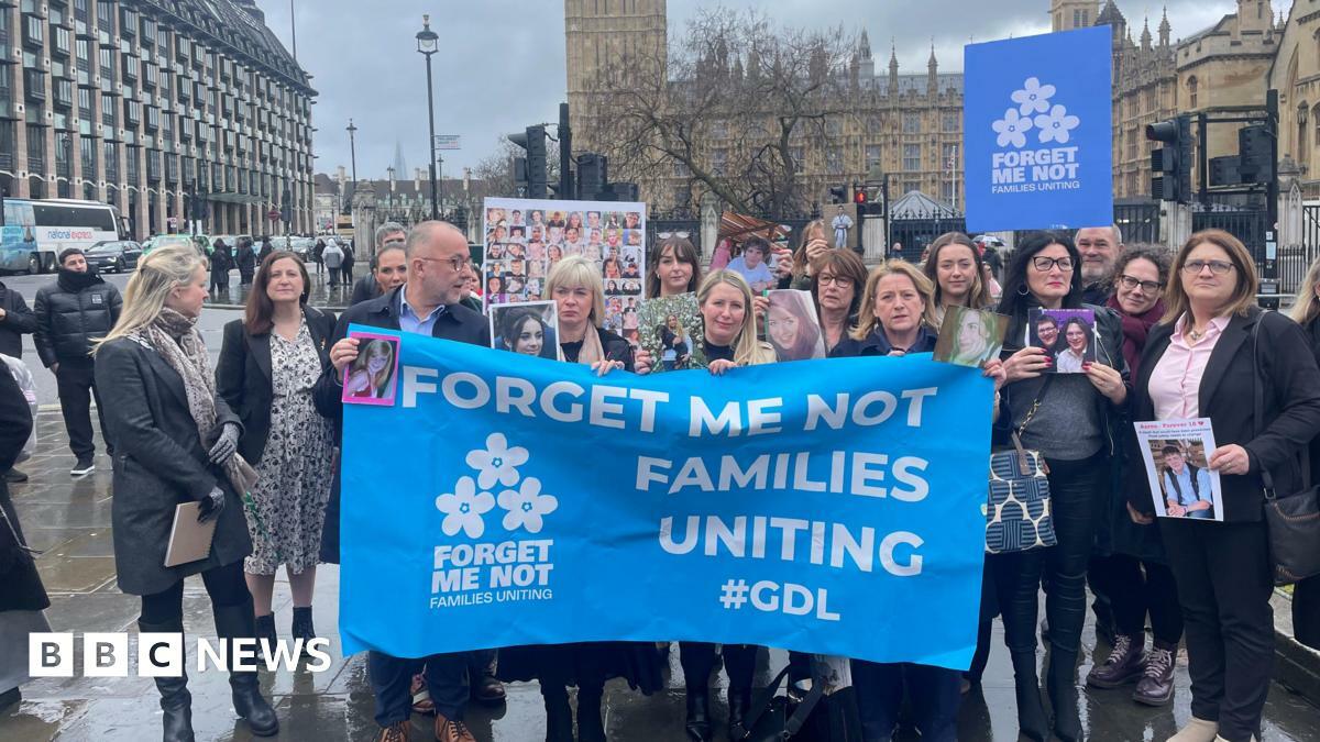A gathering of men and women with a large blue flag in front of the Houses of Parliament. The flag reads forget me not families uniting.