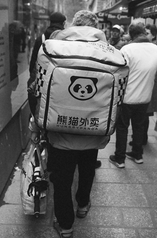 A bustling street scene captures a figure with a large "Hungry Panda" backpack, reminiscent of the iconic style of Herbert Magnin. The black and white image brings into focus the dynamic energy, with passersby blurred in the background.