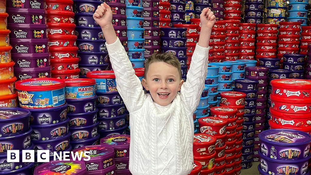 Teddy wears a white cable-knit jumper. He has short light brown hair. His arms are raised in triumph. He is in a warehouse full of thousands of sweet tubs, including Heroes, Quality Street, and Celebrations.