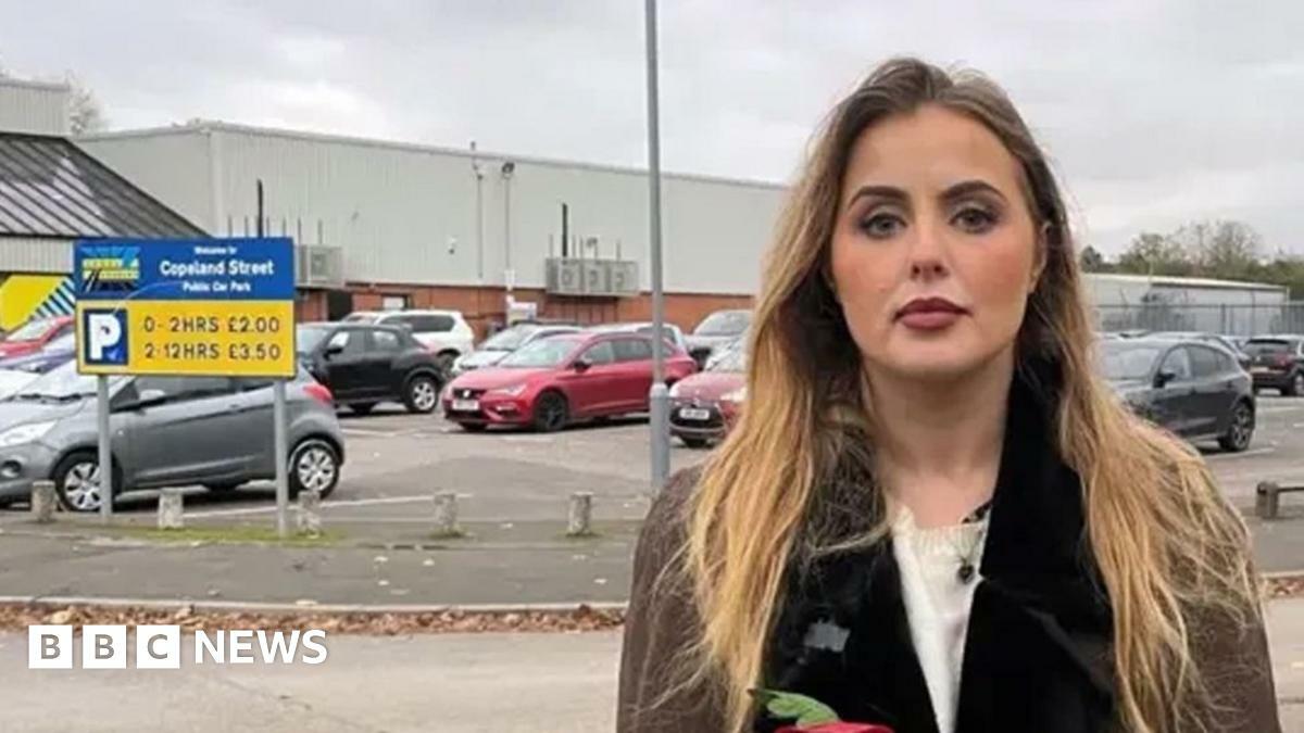 Miss Hudson, who is a young blonde woman, stands outside the Excel carpark in question near a sign showing charges, wearing a brown coat 