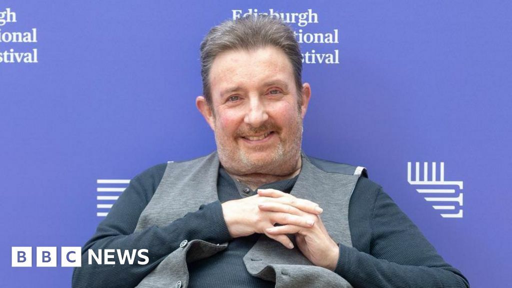 Denzil Meyrick smiles at the camera. He wears a dark grey top with a lighter grey waistcoat over the top. His hands are clasped across his chest and he is sitting down. A purple sign behind him reads Edinburgh International Festival.