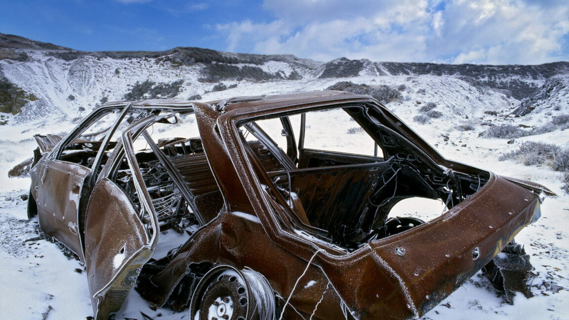 A rusted 1980s-era car wreck, with rims and pieces strewn about, in a rocky, snow-covered mountain scene.