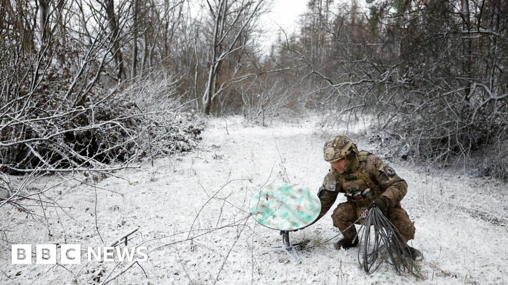 Ukrainian soldier crouches to disconnect Starlink receiver in snow-covered forest clearing on the frontline in January 2023