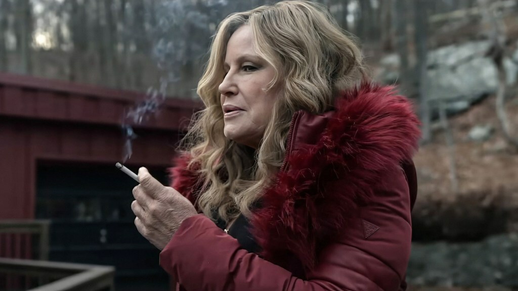 Jennifer Coolidge wears a red fur-trimmed coat and smokes a cigarette outside a house in the woods in a still from 'Riff Raff'.
