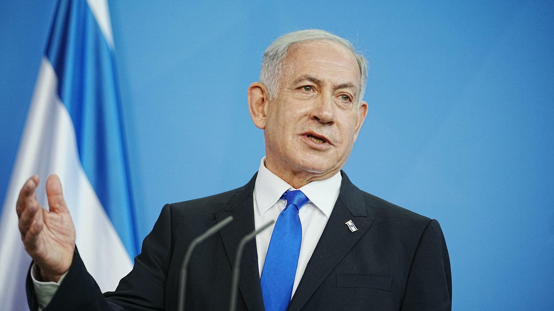 Benjamin Netanjahu, Ministerpräsident von Israel, steht vor einer blauen Wand; er trägt Anzug und Krawatte und spricht in ein Mikrofon. Dabei hebt er die rechte Hand. 