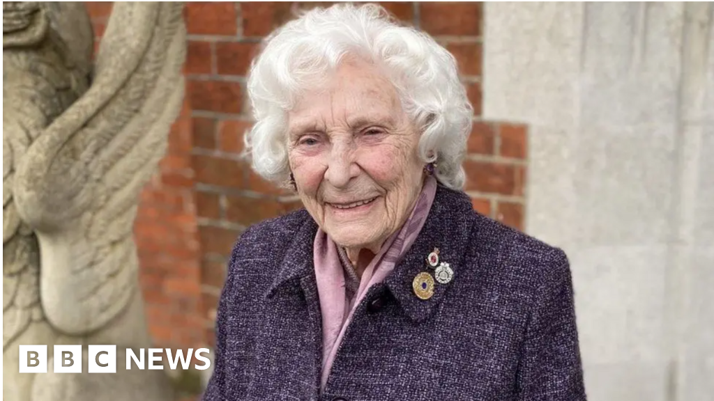 Pictured is Betty Webb with short curly white hair and in a purple coast with army pin badges on the right lapel. She is slightly smiling and looking towards the camera.