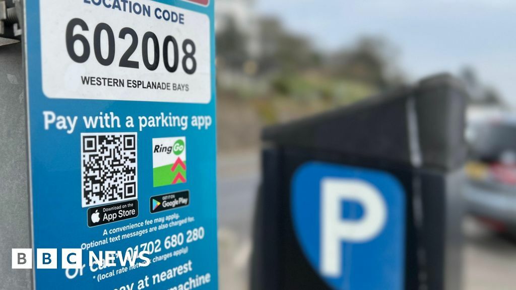 A parking meter with accompanying signage featuring a prominent QR code. The background, of a car park, is blurred.