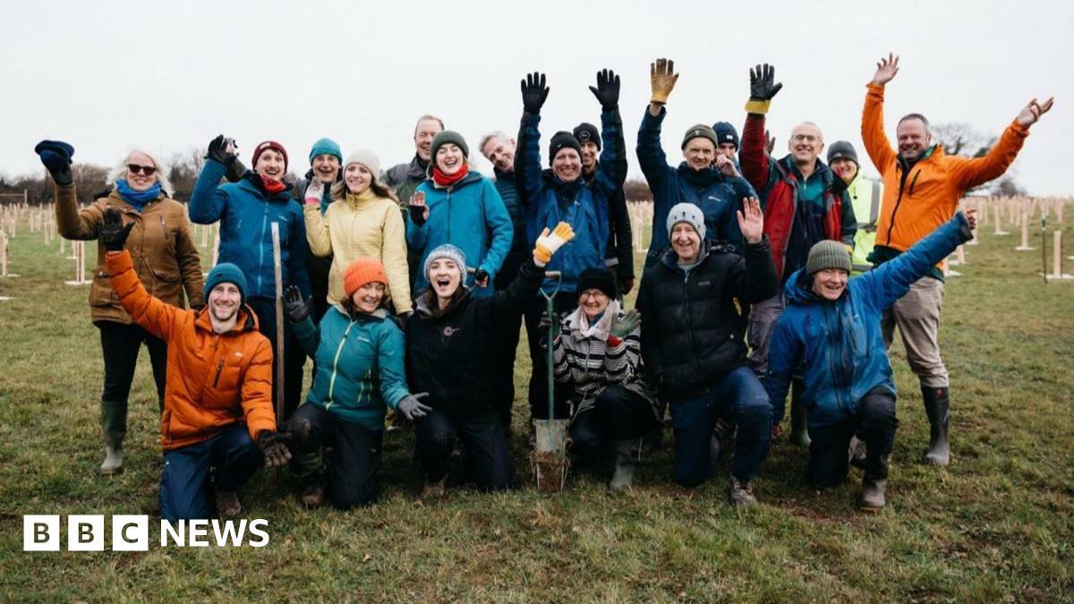A large group of volunteers wearing rain jackets, gloves and hats. They are standing in a large field surrounded by tree saplings which have just been planted, and are being held up with plastic tree guards. It is a misty, wet day but the group are smiling and waving their arms in the air.