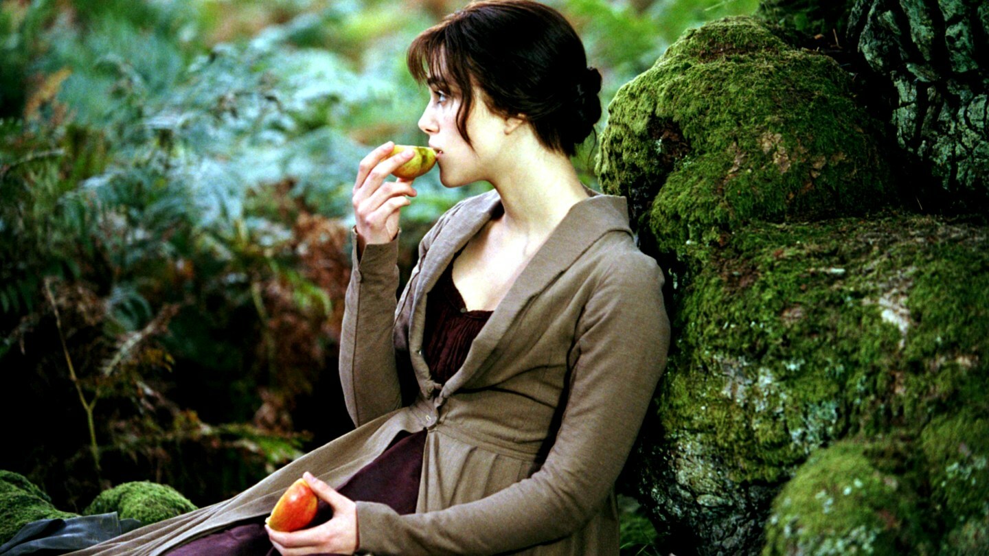 Keira Knightley eats an apple propped against a tree in a scene from 2005's 'Pride & Prejudice' while wearing an overcoat and purple dress