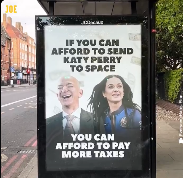 Billboard on the side of a bus stop shows a laughing Jeff Bezos and Katy Perry in her space suit.  Text reads:
IF YOU CAN AFFORD TO SEND KATY PERRY TO SPACE
YOU CAN AFFORD TO PAY MORE TAX