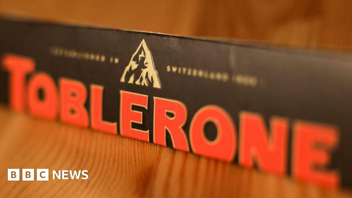 An image of a Toblerone bar on a wooden table. The bar has black packaging with 'Toblerone' written in red. 