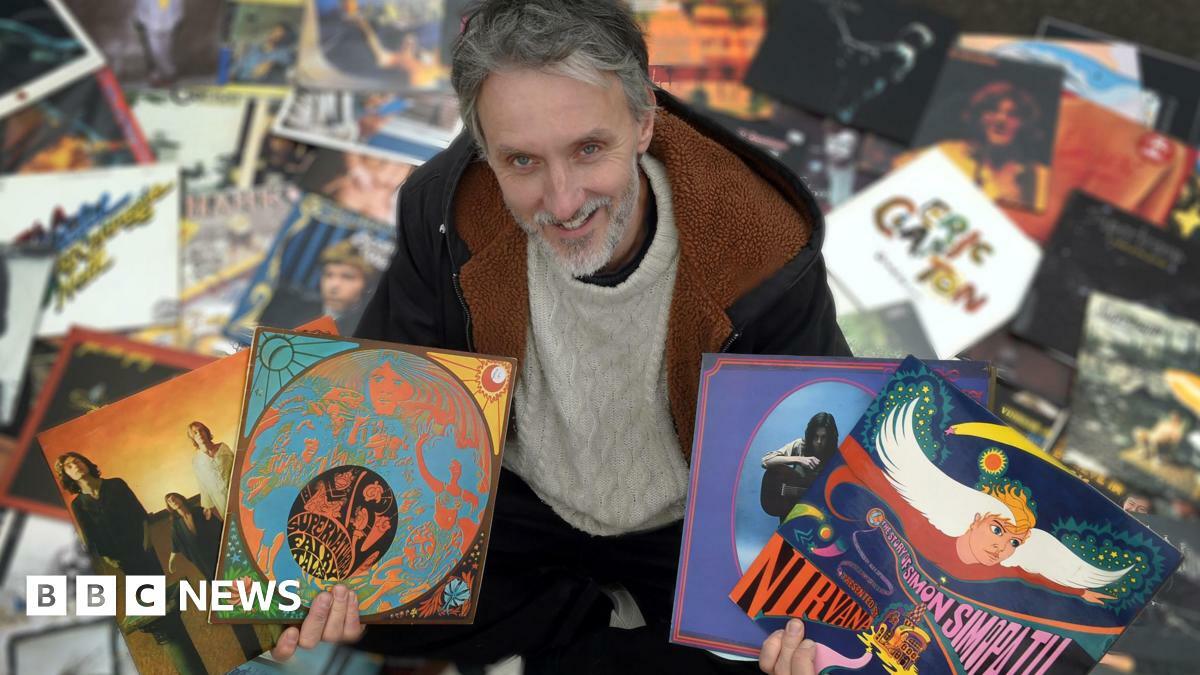 A man with grey hair and a bear standing among a number of vinyl record sleeves. He is holding four in his hands, showing the artwork on the front.