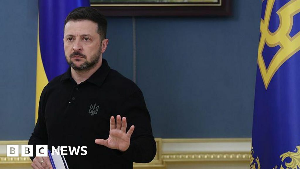 Volodymyr Zelensky speaks at a press conference, holding one hand up against a backdrop of Ukrainian flags