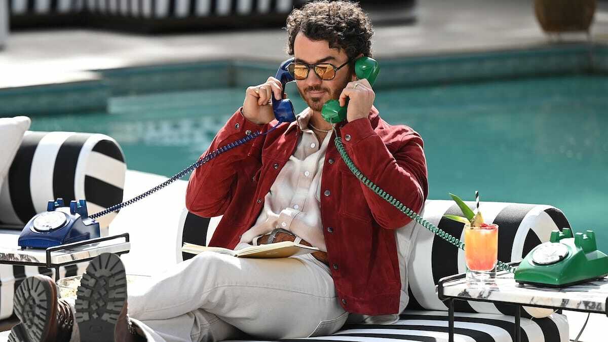A person sits on a poolside chair talking into two corded phones