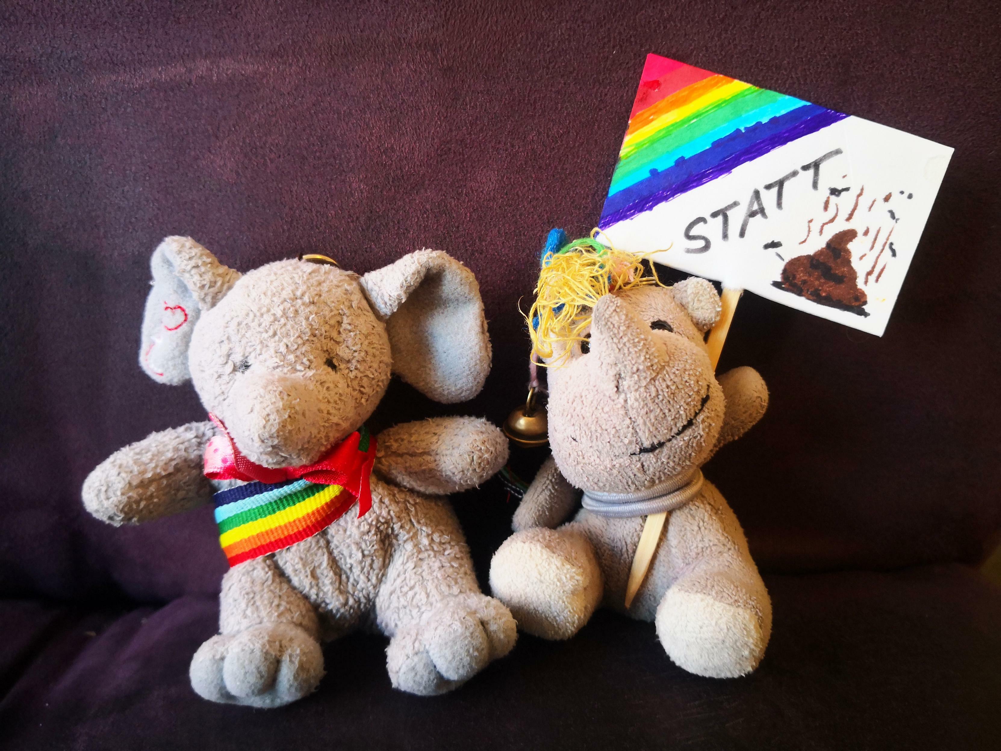 Die beiden kleinen Plüschies Elefanty Elfi, das ihr Regenbogenband schick als Schärpe trägt, und Einhorn Carola mit einem Demoschild vor der Demokratie-Demo am 20.1.2024, im Hintergrund der Stoff (Sitzfläche und Rückenlehne) des dunkelvioletten Sofas.