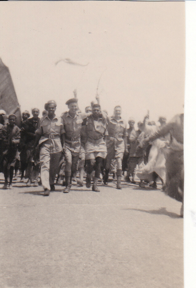 Photo of three smiling men in service uniforms striding arm-in-arm towards the camera, closely followed by another waving a flag on a stick; and a small group of Egyptian civilian men, several of them also waving.
