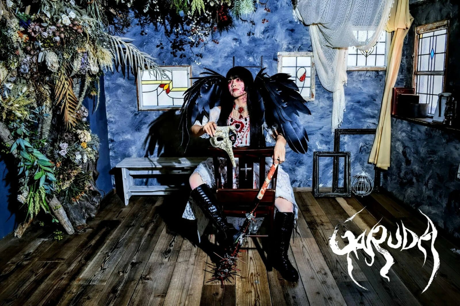 The photo shows Yuffie Sakimura as GARUDA sitting on a (prayer) chair in a room with gothic elements, plants and blue walls. She is dressed in a white dress with red elements, wearing black boots and black wings on the back, and simultaneously holding a bully mask and a baseball bat with spikes while looking away from the camera.
